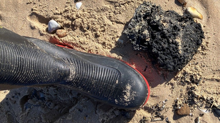 Black welly boot placed next to a charred lump of black resin on a beach
